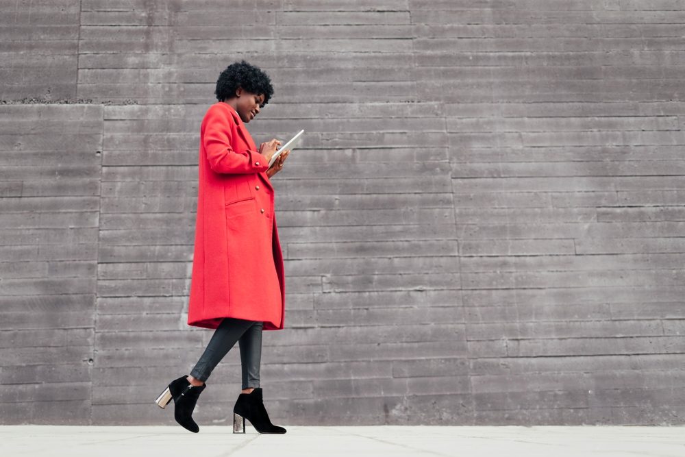 women in red coat walking while looking at i-pad. the article is about  Dallas outfit ideas.  