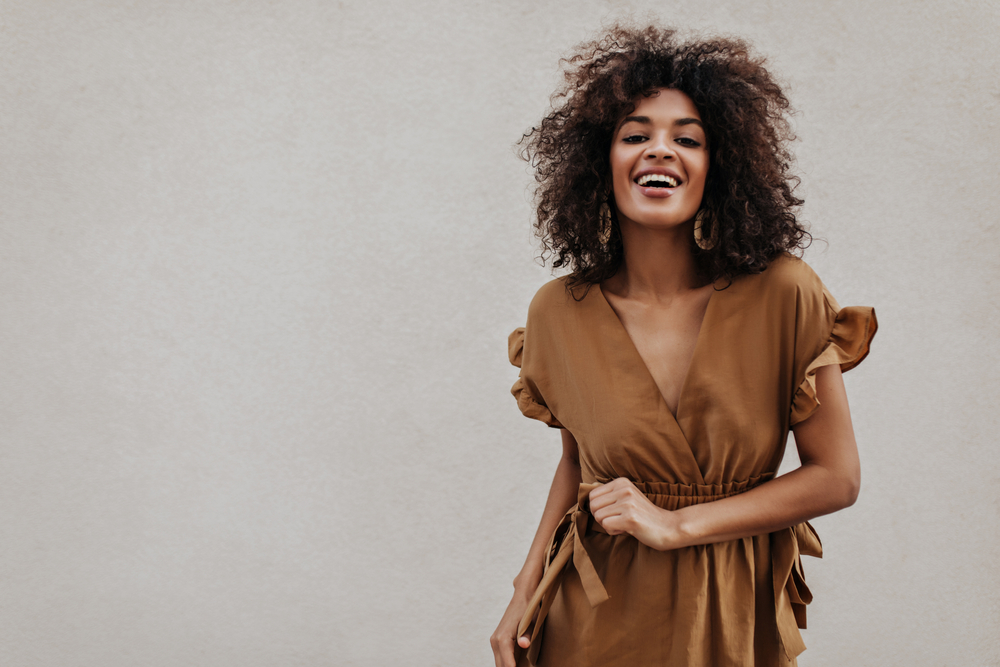 Brown-eyed woman in brown dress smiles on grey background. Stylish brunette curly dark-skinned lady