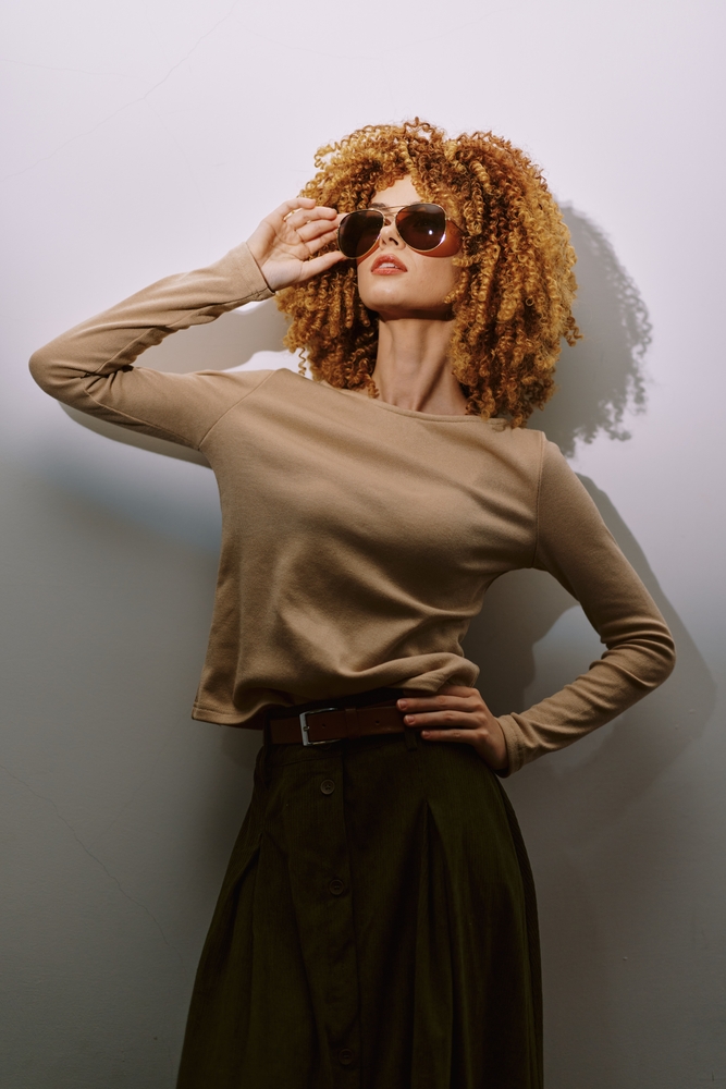 Fashion model with curly hair wearing sunglasses, posing against a light background The outfit features a neutral top and a dark skirt