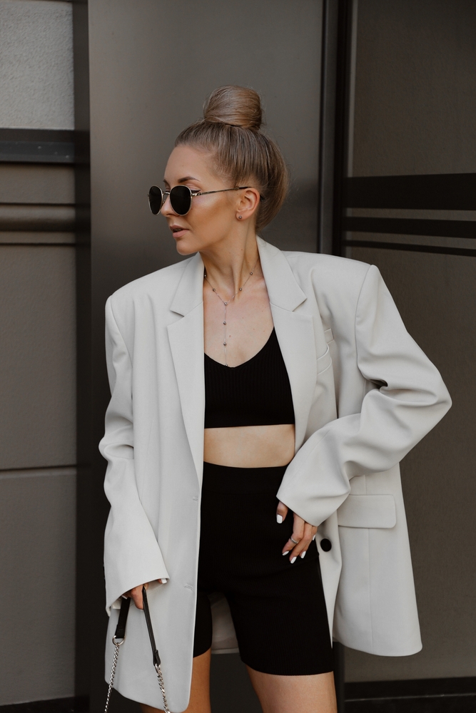 portrait of beautiful young woman dressed in beige oversized jacket, black knitted suit with shorts and top, bag, accessories, sunglasses, bun hairstyle