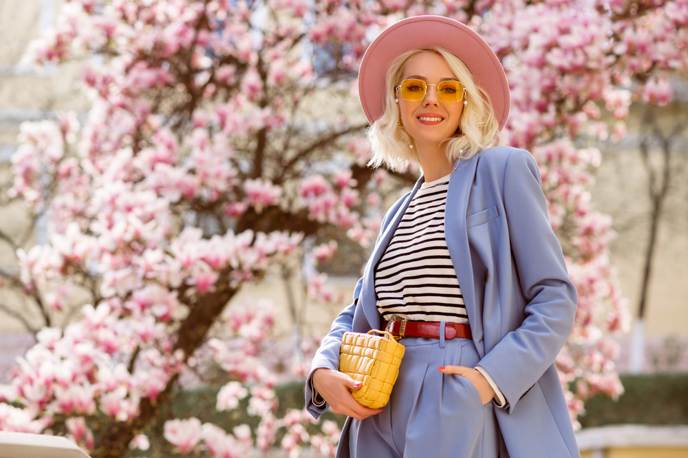 Spring fashion, street style portrait of elegant happy smiling woman wearing pink hat, yellow sunglasses, blue suit, striped t-shirt, holding trendy leather bag, posing outdoor. 