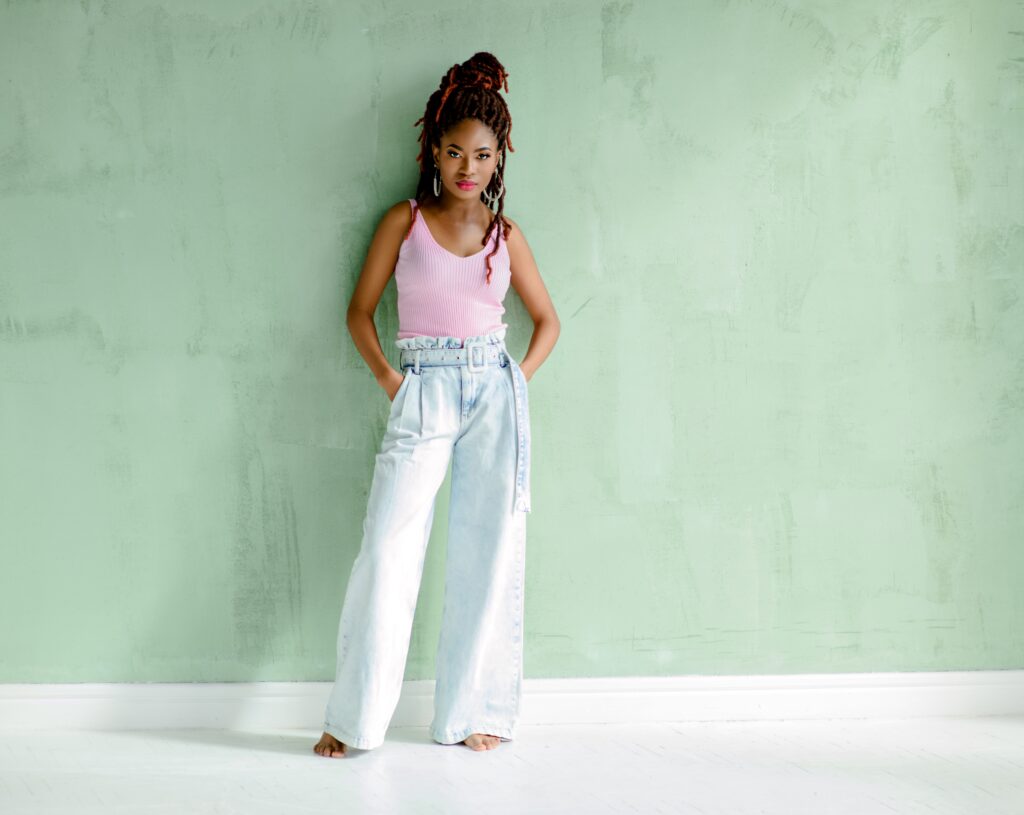 Beautiful African young woman looking at the camera, standing barefoot with her hands in pockets, isolated on a light green wall. One of the  Dallas outfit ideas