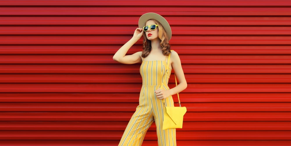 young woman model posing with handbag wearing yellow jumpsuit, summer straw hat on red background