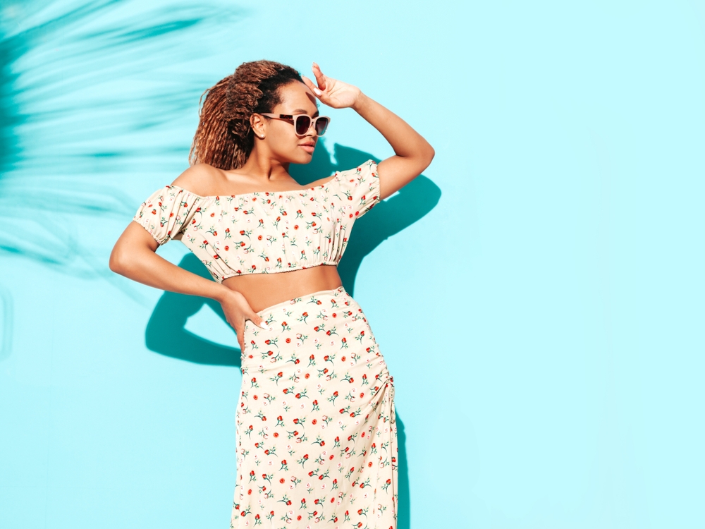 woman standing wearing a two piece top and skirt with her hand up to her face standing against a blue wall