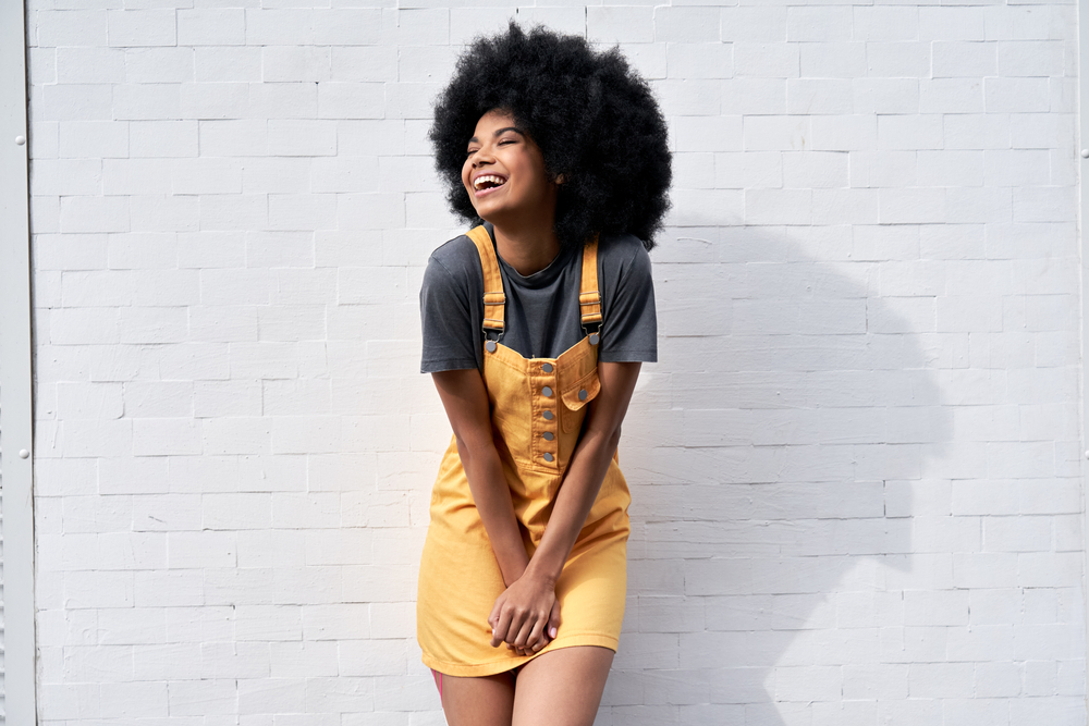  young woman wears yellow trendy sundress with Afro hair laughing looking away standing against white brick wall outdoor background.